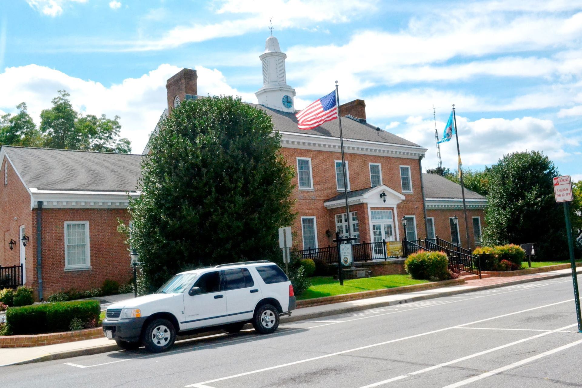 A red brick building flying  the american flag and the delaware state flag sits on the edge of a street as  a white SUV drives by. 
